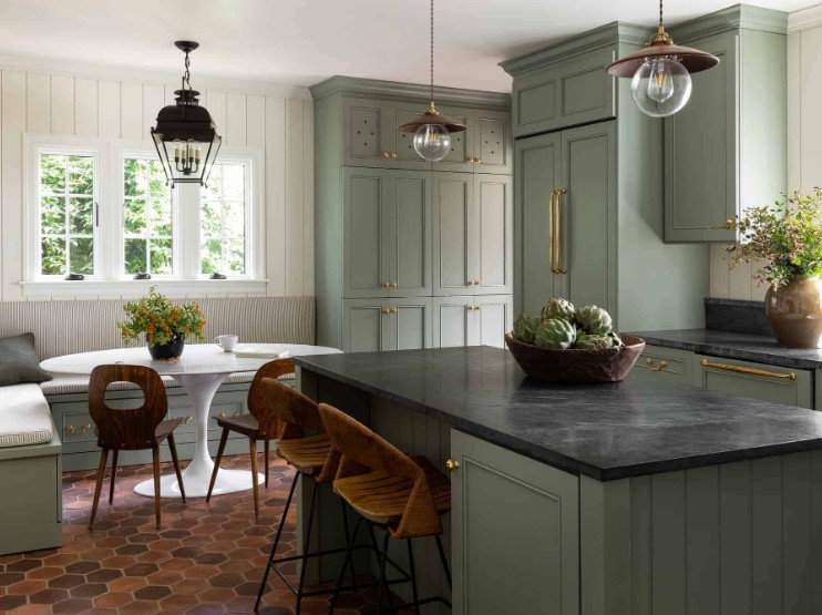 a sage green kitchen with shaker cabinets, black stone countertops, gold handles, pendant lamps and an eating zone in the corner