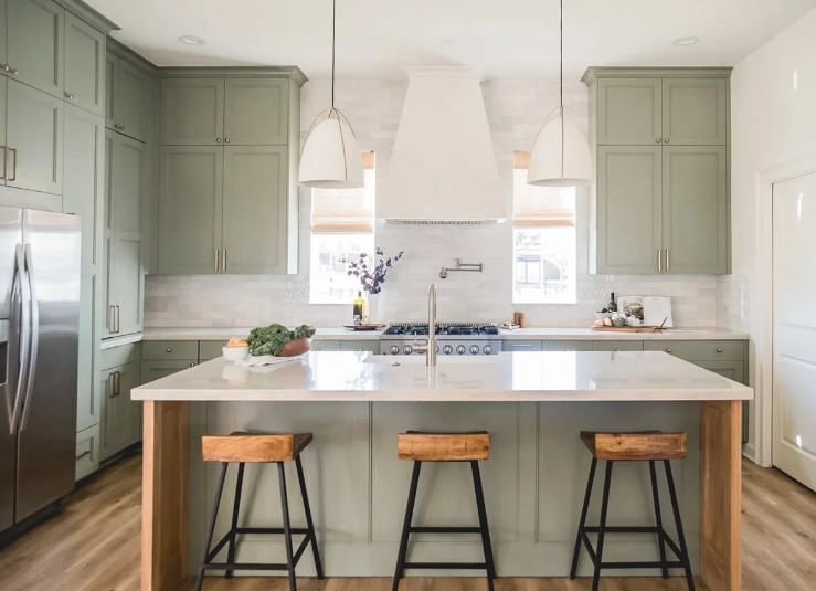 a sage green kitchen with shaker cabinets, white stone countertops and a white tile backsplash, white pendant lamps and cork stools