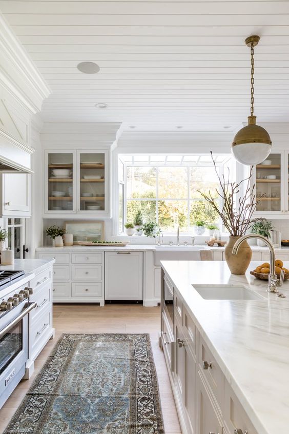 a serene white kitchen with inlay and glass cabinets, a white stone backsplash and countertops, brass pendant lamps