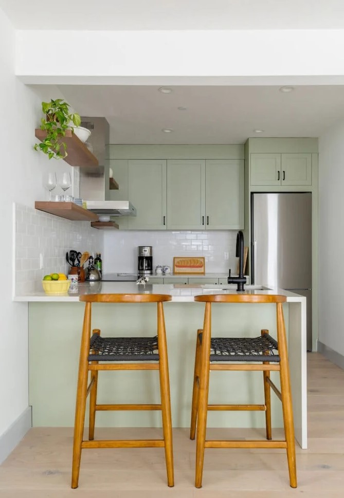 a small and cool sage green kitchen with shaker cabinets, white stone countertops and a white subway tile backsplash, black handles for a modern touch