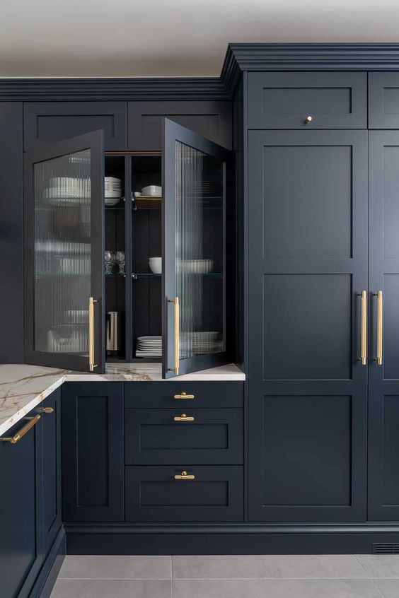 a sophisticated midnight blue kitchen with shaker cabinets and a fluted glass one, with gold fixtures and a white stone countertop