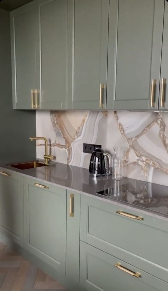 a sophisticated sage green kitchen with shaker cabinets, a beautiful marble backsplash and sleek countertops plus gold handles