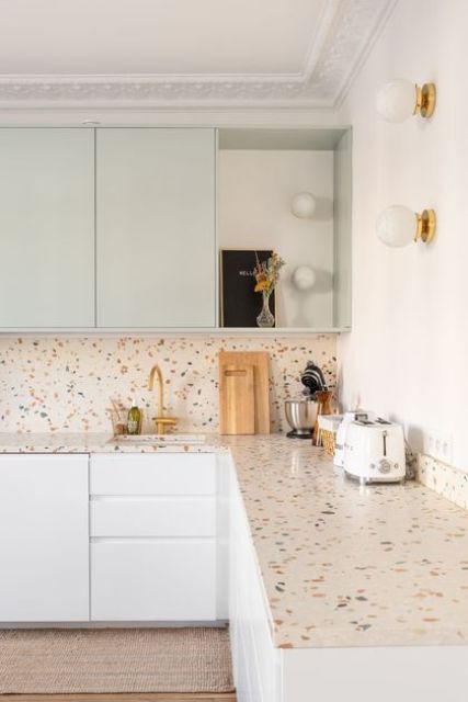 a sutble kitchen with white and light green flat panel cabinets, a colorful terrazzo countertop and a backsplash, touches of gold