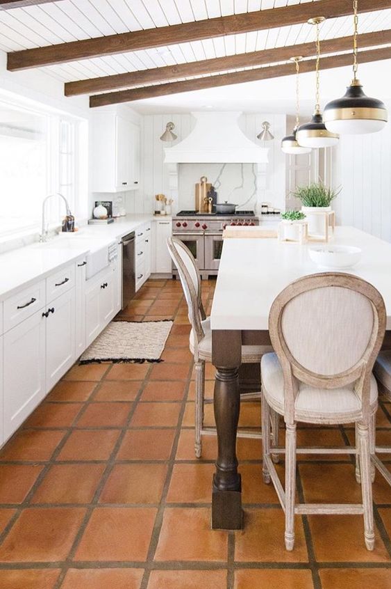 a vintage-inspired white kitchen with only lower cabients, a large cooker and a hood, stained beams, a large table that is a kitchen island