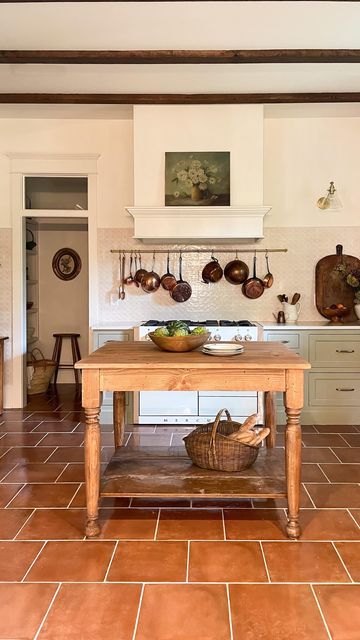 a vintage kitchen with sage green shaker style cabinets, a large hood, a glossy tile backsplash, a wooden table and some artworks