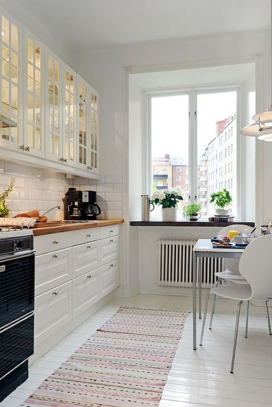 a welcoming white Scandinavian kitchen with glass cabinets, white tiles, butcherblock countertops and a small eating zone