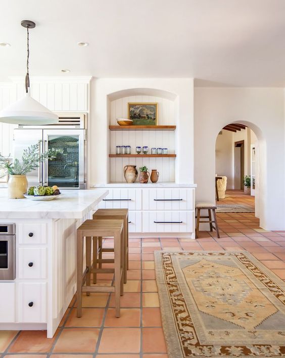 a white famrhouse kitchen with a terracotta tile floor, white shiplap cabinets, niches, a large kitchen island and pendant lamps