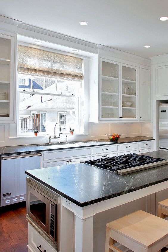 a white farmhouse kitchen with shaker cabinets, black soapstone countertops, a window as a backsplash