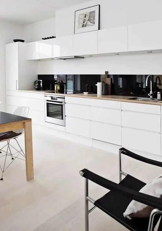 a white minimalist one wall kitchen with blonde butcherblock countertops, a black glass backsplash and an eating zone