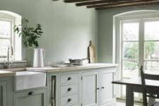 an airy sage green kitchen with shaker cabinets, butcherblock countertops, sage green walls, stained wooden beams and a table