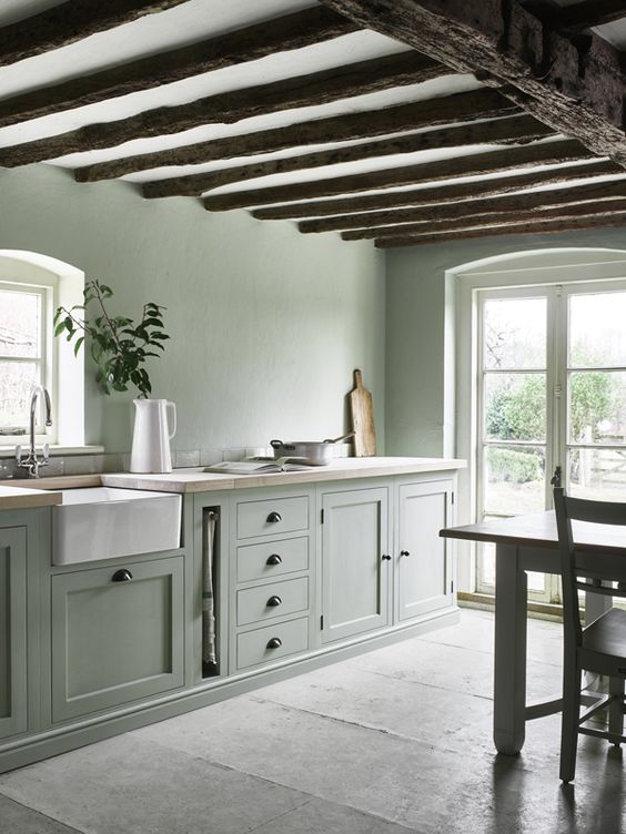 an airy sage green kitchen with shaker cabinets, butcherblock countertops, sage green walls, stained wooden beams and a table