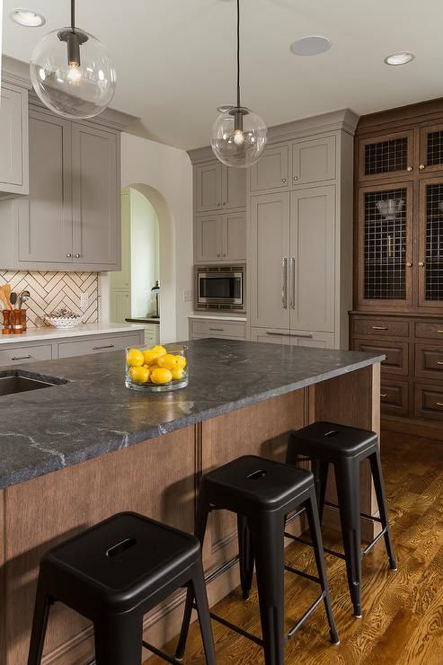 an elegant kitchen with grey shaker style cabinets, a white herringbone tile backsplash, a stained kitchen island with a soapstone backsplash