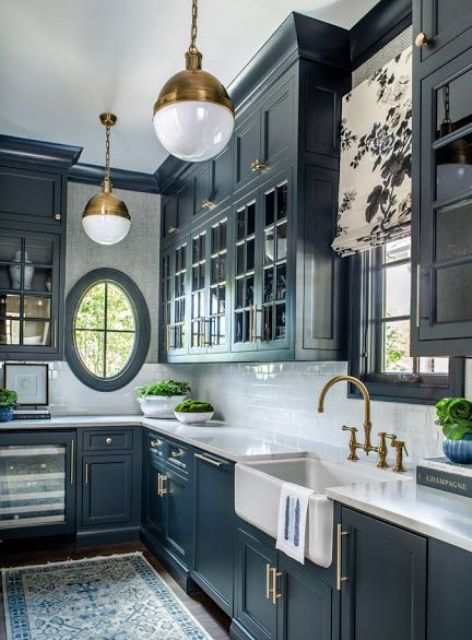 an elegant navy kitchen with shaker and glass front cabinets, a white tile backsplash, an oval window, brass pendant lamps