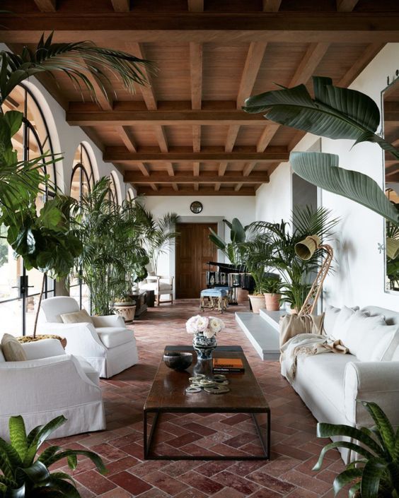 an outdoor living room with a terracotta tile floor, white seating furniture, a low coffee table, potted plants and a wooden ceiling with beams