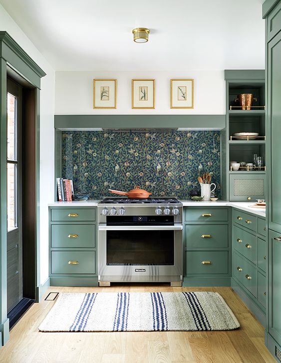 a beautiful green kitchen with flat panel cabinets, a dark floral wallpaper backsplash covered with glass and a built-in hood