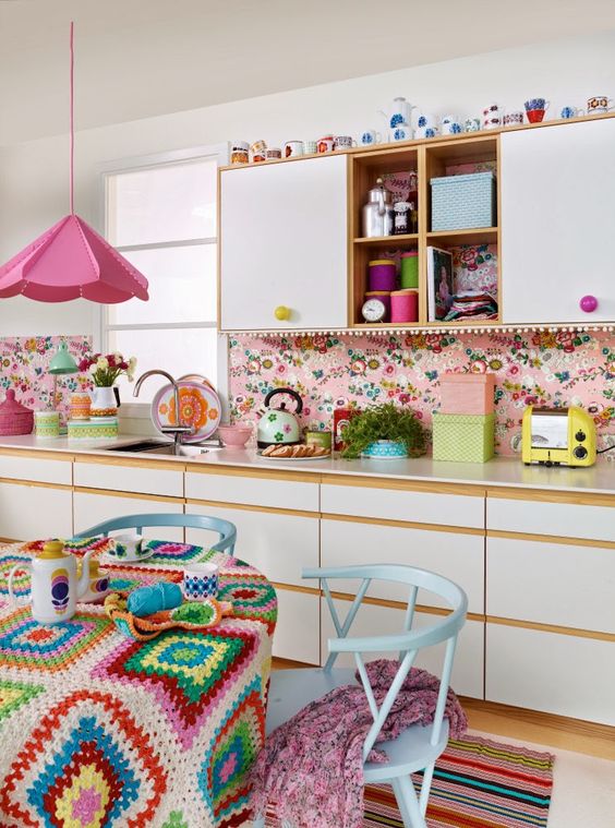 a bright retro kitchen with white flat panel cabinets, a bright pink wallpaper backsplash, a bright pink pendant lamp and pastel blue dining furniture