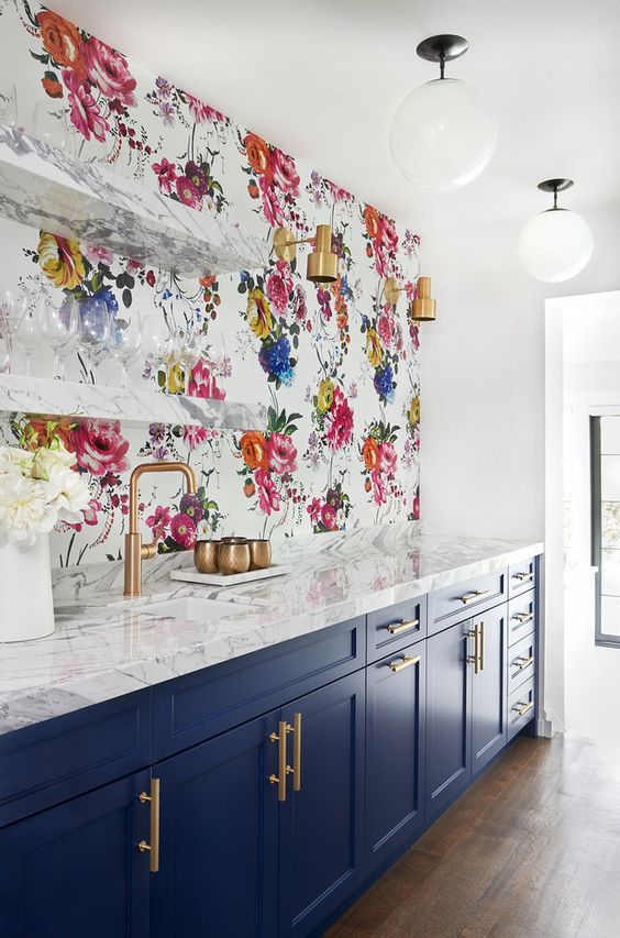 a navy and white kitchen with shaker cabinets, white marble countertops, a bright floral wallpaper wall and open shelves plus brass touches
