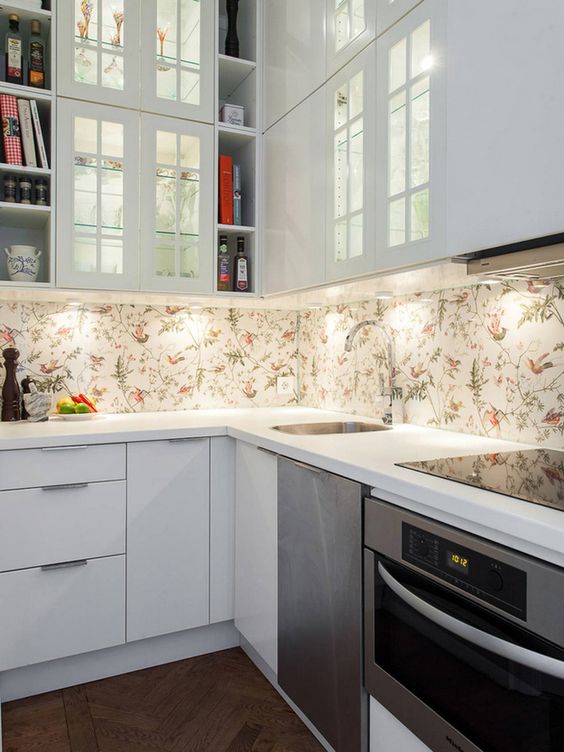 a small white kitchen with flat panel cabinets, a floral wallpaper backsplash and glass front cabinets plus sleek stone countertops