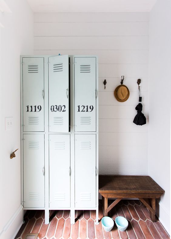 a small and cool mudroom with a terracotta tile floor, pale green lockers, a dark-stained bench and some necessary stuff