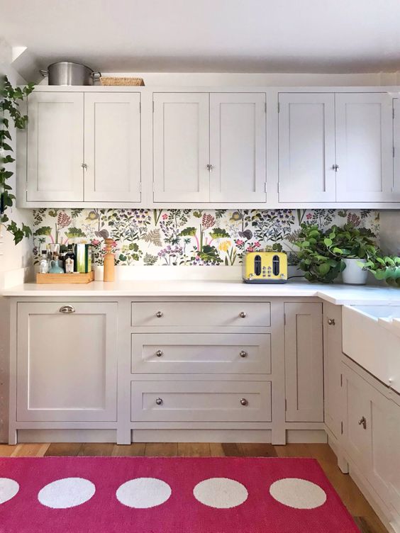 a white kitchen accented with a bright floral wallpaper backsplash, white countertops and potted plants plus a bold polka dot rug