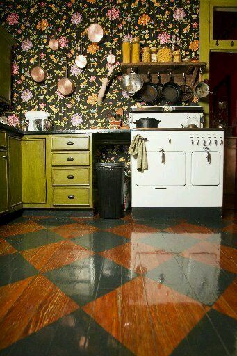 an eclectic kitchen with green cabinets, dark floral wallpaper, a vintahe hearth and some pretty dishes plus a bold parquet floor