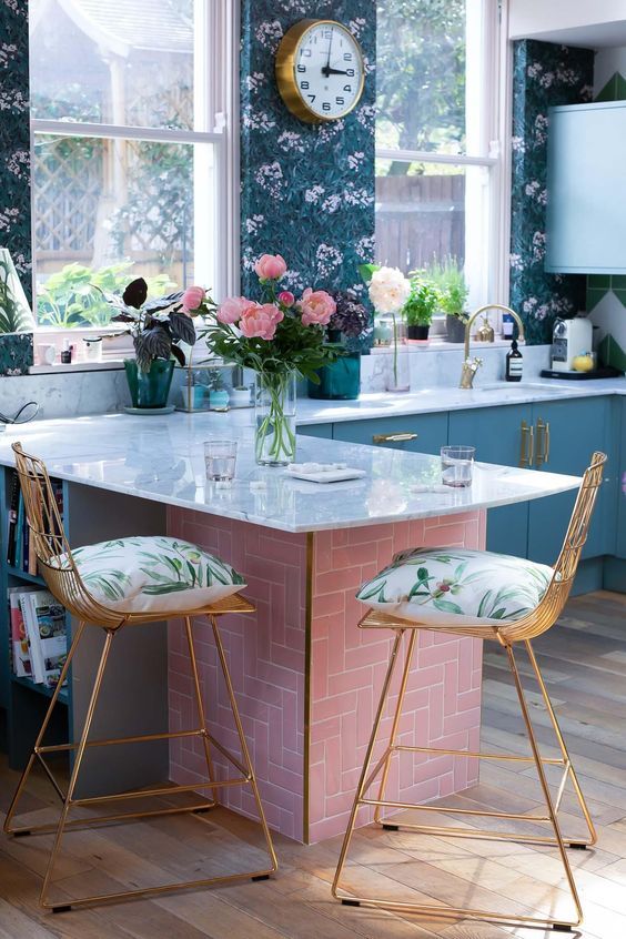 a blue kitchen with flat panel cabinets, bright blue and green floral wallpaper, white countertops and gold stools with bright pillows