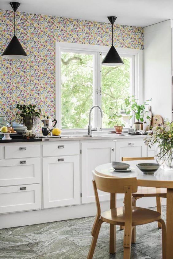 a chic cottage kitchen with white shaker cabinets, a bright floral wallpaper wall, stone countertops and black pendant lamps