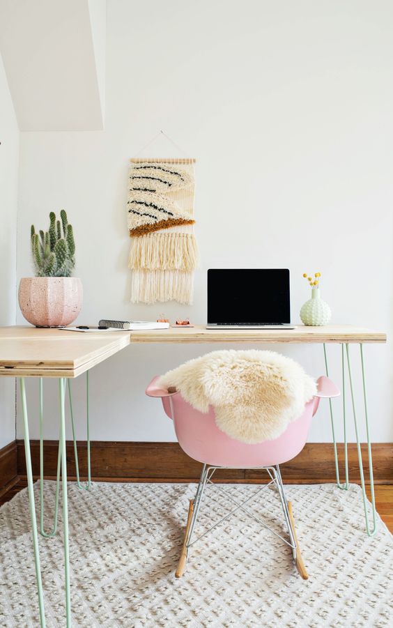 a lightweight corner desk with light green hairpin legs, a pink chair, a boho rug, a macrame piece and a potted plant