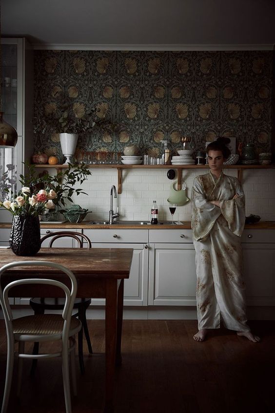 a chic creamy kitchen with shaker cabinets, butcherblock countertops, an open shelf and dark floral wallpaper plus some decor