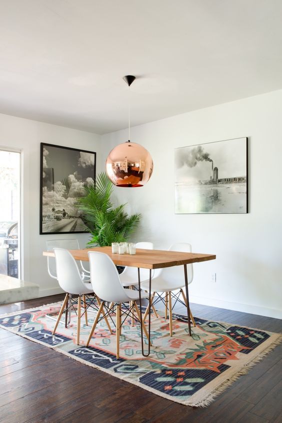 a mid-century modern dining room with a hairpin leg dining table, white chairs, a printed rug, a copper pendant lamp and some art