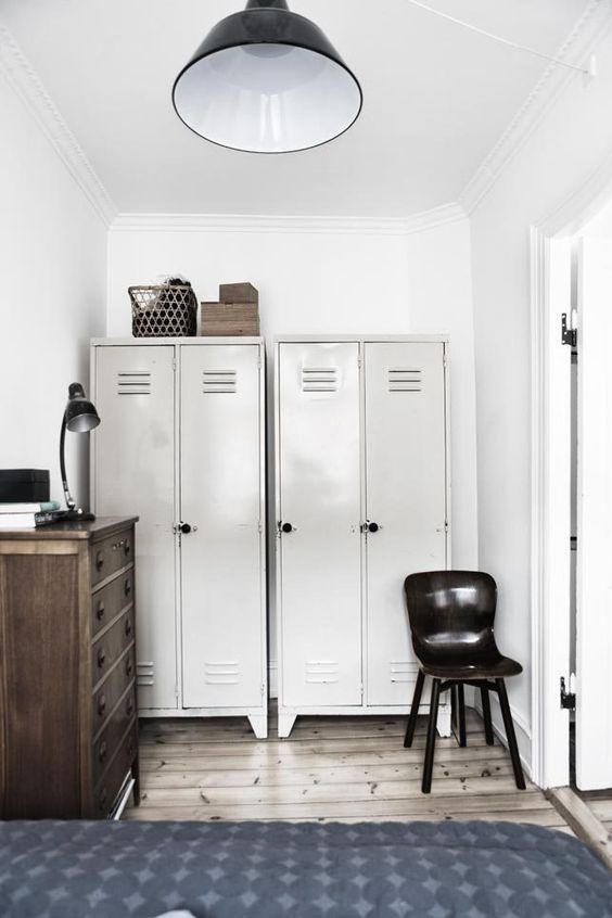 a Nordic bedroom with greyish lockers fo storage, a dark-stained dresser, a bed, a black chair and black metal lamps