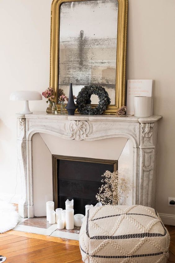 a French fireplace with a decorated mantel, an arrangement of candles, a boho pouf and a mirror in a gilded frame