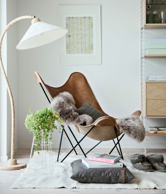 a Scandinavian nook with a brown leather butterfly chair, a neutral rug and a pillow, a floor lamp and a trellis with greenery