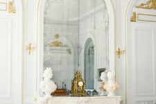 a beautiful French fireplace with decor on the mantel and an arched mirror, gold decor and details here and there