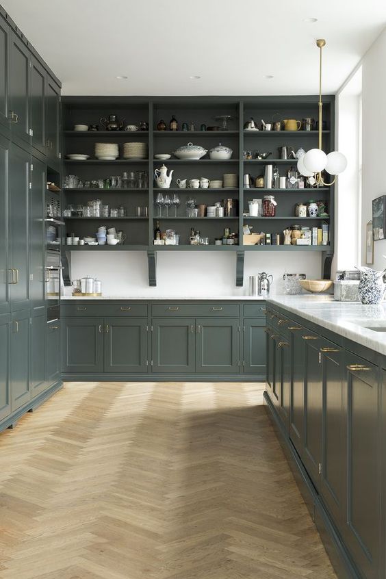 a beautiful dark green English-inspired kitchen with shaker and open cabinets, white countertops and a backsplash, brass and gold fixtures