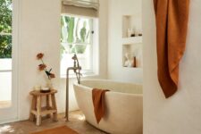 a beautiful earthy bathroom with white plaster walls, a parquet flooring, a stone bathtub, niches with decor and rust textiles