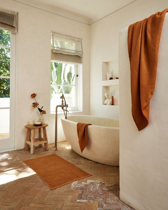 a beautiful earthy bathroom with white plaster walls, a parquet flooring, a stone bathtub, niches with decor and rust textiles