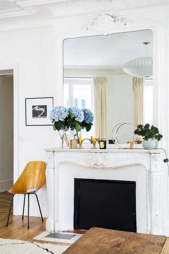 a beautiful white ornated Franch fireplace with an oversized mirror on top, a plywood chair and a wooden coffee table