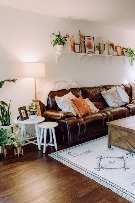 a boho living room with a brown leather sofa, a wooden coffee table, side tables, a shelf with art and lots of greenery