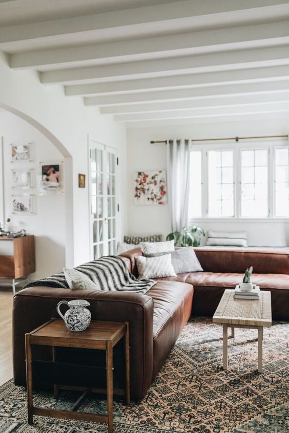 a boho rustic interior with a brown leather sectional, a coffee table and a side one, a large printed rug and pillows