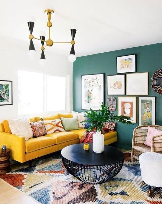 a bright boho living room with a green wall and a gallery wall, a yellow velvet sectional, a retro chandelier and a black round table