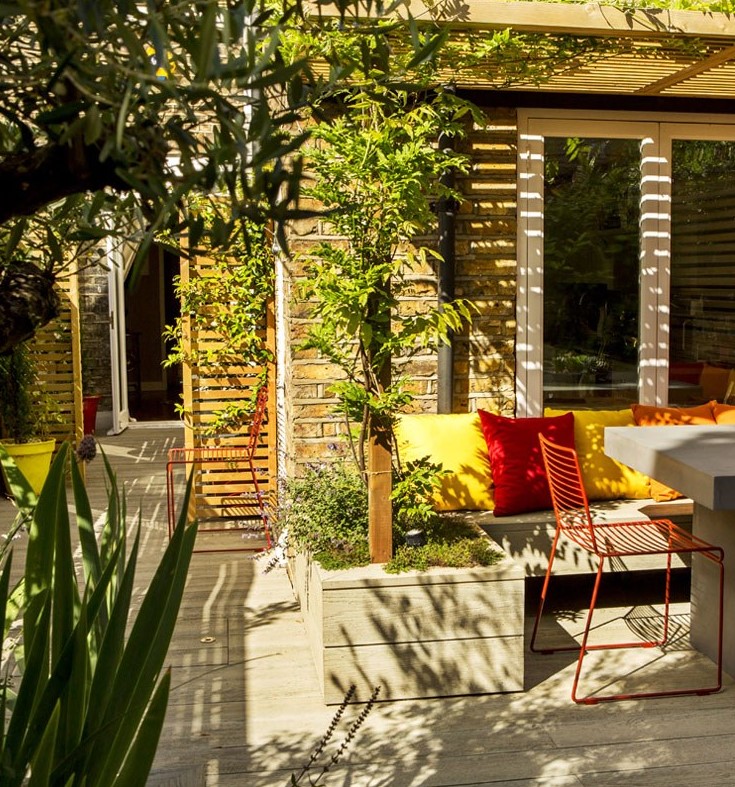 a bright outdoor space with limed oak decking, a bench with bold pillows, potted greenery and plants, red metal chairs