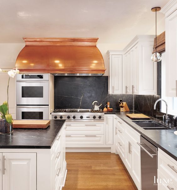 a catchy and chic creamy kitchen with a vintage feel, black soapstone cabinets and a backsplash, a copper hood and stainless steel appliances