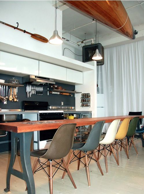 a catchy modern industrial kitchen with white and metal cabinets, a black backsplash, a long table and colorful Eames chairs