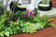 a catchy outdoor space with millboard decking, an outdoor fireplace, black woven chairs with green upholstery, grasses, greenery and blooms plus a tree over the space
