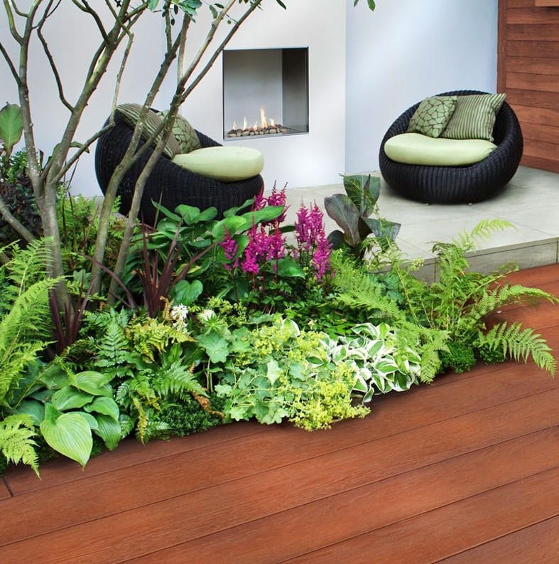 a catchy outdoor space with millboard decking, an outdoor fireplace, black woven chairs with green upholstery, grasses, greenery and blooms plus a tree over the space