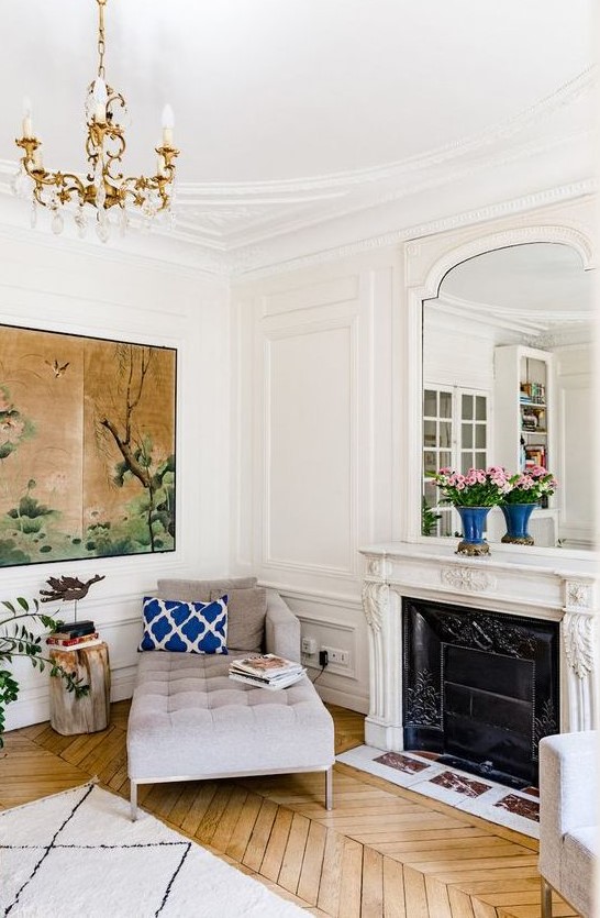 a chic Parisian nook with a grey daybed, a French fireplace and a mirror over it, a parquet floor, an oversized artwork