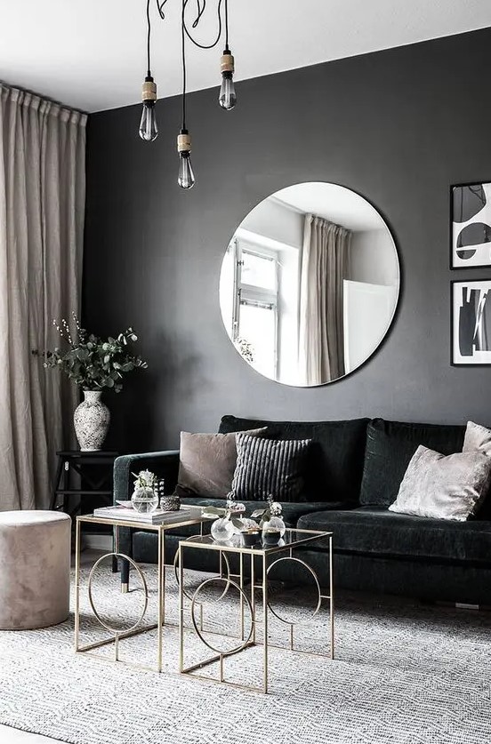 a chic and dramatic living room with a black accent wall, a black velvet sofa, chic glass coffee tables, pendant bulbs and a round mirror