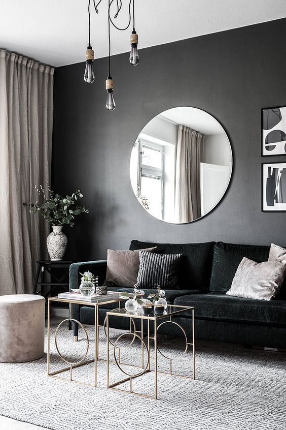 a chic and dramatic living room with a black accent wall, a black velvet sofa, chic glass coffee tables, pendant bulbs and a round mirror