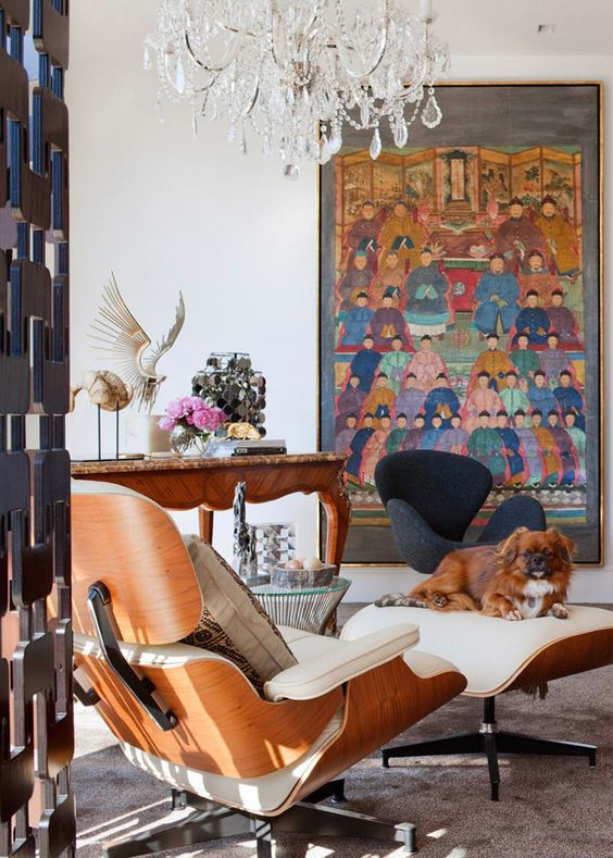 a chic and eclectic space with a bold artwork, a creamy Eames lounger and ottoman, a carved desk and a crystal chandelier